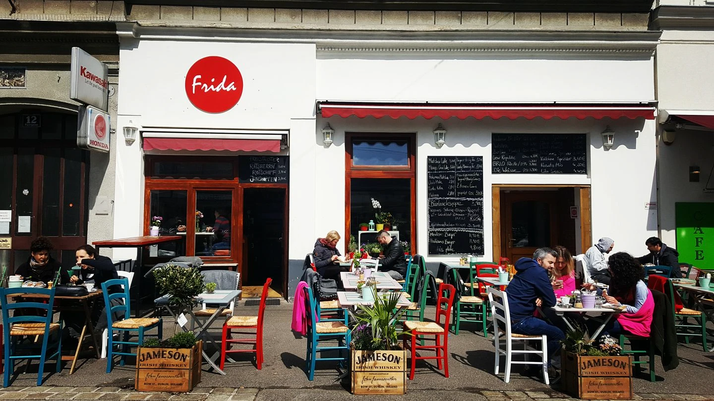 Gäste sitzen im sonnigen Außenbereich des Cafés Frida und genießen Getränke und Speisen an bunten Tischen.